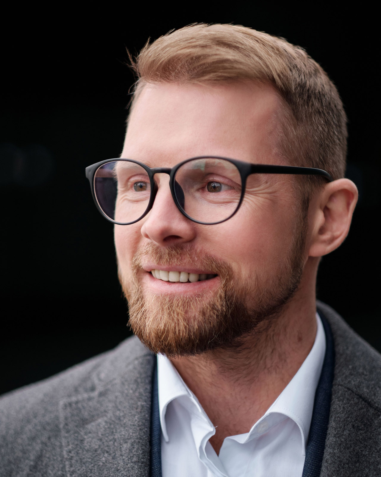 A man with glasses and a light beard wearing a gray blazer over a white shirt, smiling warmly